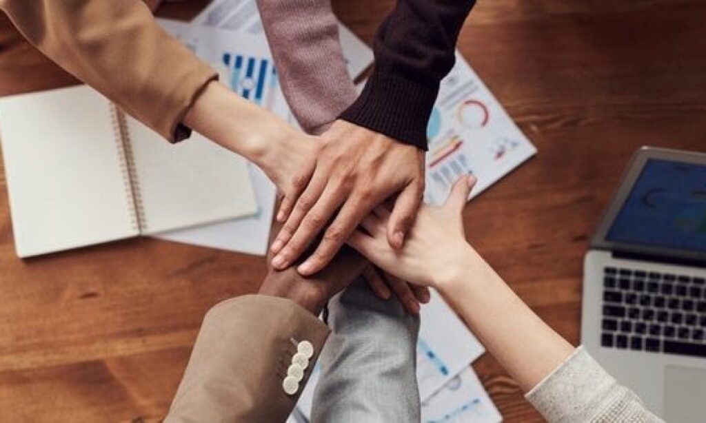 People placing their hands together over a table, symbolizing teamwork, social cohesion, unity, and collective progress.
